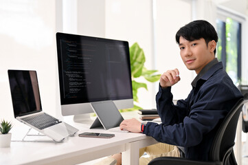 Smart Asian male web developer sits at his modern office desk, smiling and looking at the camera.