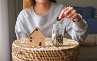 Wall Mural - Closeup image of a woman putting coins into a glass jar with wooden house models for saving money concept