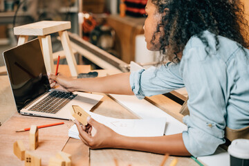 Wall Mural - Carpenter america black woman curly hair sketch making notes in work paper while standing at wooden table with laptop computer, young female working learning online at woodshop, Happy Carpenters Day