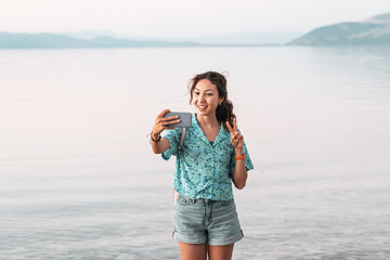 Wall Mural - Happy girl taking selfie photo on a high cliff with splendid view of panoramic seascape or lakeshore and mountains.