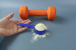 A woman's calloused hand holds a portion of whey protein in front of a dumbbell. The concept is to replenish the necessary amount of protein consumed by the athlete.