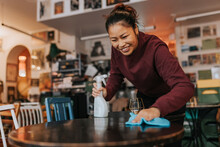 Happy Female Coffee Shop Owner With Spray Bottle And Rag Cleaning Table In Cafe