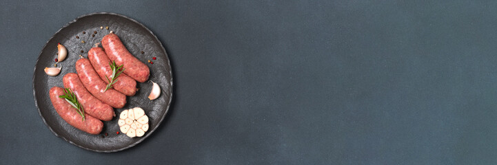 Long banner with raw sausages or bratwurst with seasonings on black plate over black stone background with copy space. Traditional pork sausages ready to cook. Overhead view