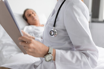 Wall Mural - Doctor in white coat holding paper folder near blurred patient in hospital ward.