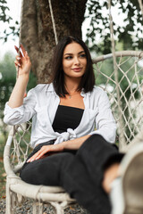 Wall Mural - Beautiful happy girl in fashionable casual clothes with a white shirt sits and rides on a knitted vintage swing in the park