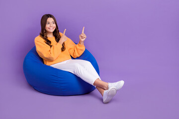 Sticker - Full length photo of dreamy cute girl dressed orange sweatshirt bean bag pointing empty space isolated purple color background