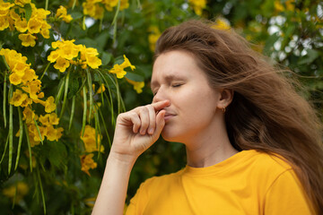 Portrait of beautiful young allergic woman is suffering from pollen allergy or cold on natural flower flowering tree background at spring or sunny summer day sneezes, blowing her runny nose rubs eyes