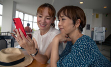 Two Mature Japanese Women, Friends Side By Side, Sitting Looking At A Mobile Phone Screen.