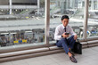 © Mint Images - A young businessman in the city, on the move, sitting on a walkway, looking at his phone.
