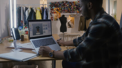 African American fashion designer develops clothes design in 3D modeling program on laptop using digital tablet and stylus. He works in atelier workshop. Fashion and technologies in business concept.