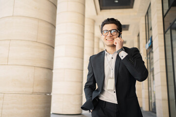 Wall Mural - A confident businessman in a formal suit on the way to the office talks to a partner on the Internet using a smartphone. Education, business, the concept of an office employee.