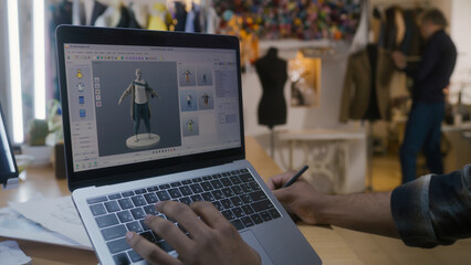 African American fashion designer creates clothes in 3D modeling software on laptop using digital tablet and stylus. He works in atelier workshop. Concept of fashion and technologies in business.
