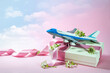 Toy airplane on a gift box with ribbon and some flowers, pink background fading into a cloudy blue sky, travel and journey present for Valentines day or wedding trip, copy space