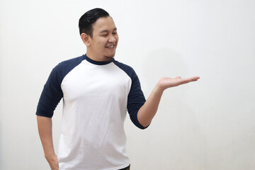 Canvas Print - Young Asian man wearing white shirt smiling, showing and pointing something on his side, against white wall with copy space