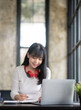 © crizzystudio - Happy Asian girl student wear wireless headphone study online with the teacher, happy young woman learn language listen lecture watch webinar write notes look at laptop sit in cafe, distance education