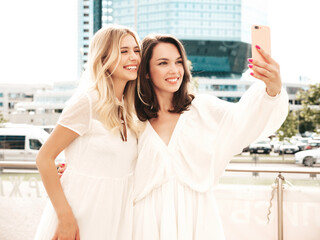 Wall Mural - Two young beautiful smiling female in trendy summer white dress clothes. Sexy carefree women walking in street. Positive models having fun, hugging and laughing. Cheerful and happy. Taking selfie