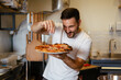 © zorandim75 - Male chef is sprinkling fresh oregano over a traditionally made home pizza.