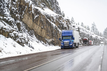 Convoy of the big rigs semi trucks with semi trailers standing on the wet road shoulder on mountain pass at winter snowy weather