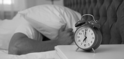 Canvas Print - Alarm clock and man covering head with pillow in bedroom, selective focus. Black and white photography