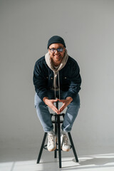 Wall Mural - Model in hat and glasses is sitting on a chair. Handsome man is in the studio against white background