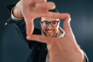 Wall Mural - Making the photo by hands, pretending. Smiling. Handsome man is in the studio against blue background