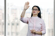 Young asian business woman is touching invisible virtual screen by her hand. Blurred windows background.