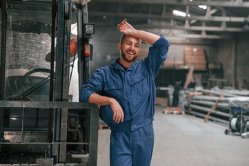 Wall Mural - Leaning on the forklift. Factory worker in blue uniform is indoors