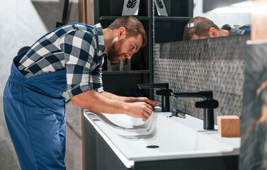 Side view. Fixing water tap. Plumber in blue uniform is at work in the bathroom