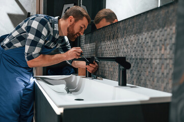 Wall Mural - Side view. Fixing water tap. Plumber in blue uniform is at work in the bathroom