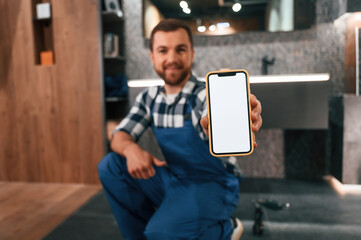 Wall Mural - Holding smartphone. Plumber in blue uniform is at work in the bathroom