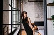 © Iona - Attractive elegant young brunette woman in black dress standing at luxurious apartment looks aside. Slim Italian girl with long hair traveling, enjoys modern hotel room. Healthy life. Successful women