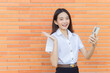© nut_foto - Portrait of beautiful an Asian Thai girl student in uniform is standing smiling happily and confidently successful while holds smartphone with brick walls as a background in university.
