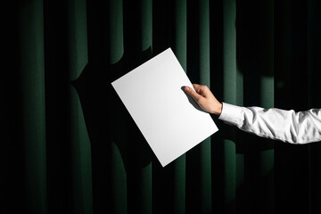 Hand in white shirt holding blank a format letter paper mockup template on a green fabric curtains background. Real photo, the isolated surface to place your design. 