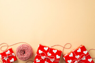 Wall Mural - Valentine's Day concept. Top view photo of gift boxes in wrapping paper with heart pattern and spool of twine on isolated pastel beige background with empty space