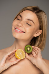 Wall Mural - joyful woman with closed eyes holding kiwi fruit and lemon isolated on grey.