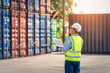 © nuttawutnuy - Engineer or foreman holding laptop and wears PPE checking container storage with cargo container background at sunset. Logistics global import or export shipping industrial concept.