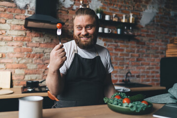 Happy guy eating salad with tomatoes at table in kitchen