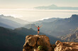 © RooM The Agency - Rear view of a woman standing on rocks with Roque Nublo natural park and Mount Teide in the background, Gran Canaria, Canary Islands, Spain