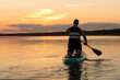 A man in shorts with his back to the camera on his knees on a SUP board with a paddle against the background of the sunset sky swims in the lake in the evening.