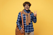 Young Cheerful Happy Teen Indian Boy IT Student He Wear Casual Clothes Shirt Glasses Bag Showing Victory Sign Gesture Isolated On Plain Yellow Color Background. High School University College Concept.