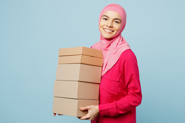 Young smiling happy arabian muslim woman wear pink abaya hijab hold stack cardboard blank boxes isolated on plain pastel light blue cyan background studio portrait. People uae islam religious concept.