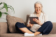 © Artem Varnitsin - Smiling senior woman sitting with crossed legs using a digital tablet. Aged female in eyeglasses reading from a digital tablet at home.