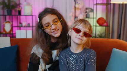 Wall Mural - Portrait of happy smiling teenage child and little sister kid looking at camera wearing stylish sunglasses. Female siblings children or best friends sitting on sofa at home playroom. Friendship family