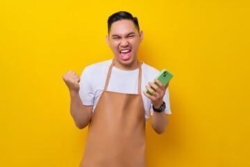 Wall Mural - excited young Asian man barista barman employee wearing brown apron work in coffee shop holding mobile phone and showing winner gesture isolated on yellow background. Small business startup concept