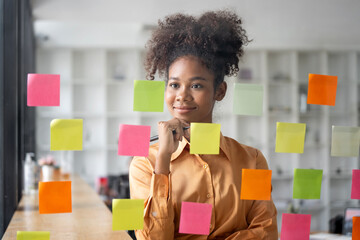 Wall Mural - Young smiley attractive african businesswoman using sticky notes in glass wall to writing strategy business plan to development grow to success