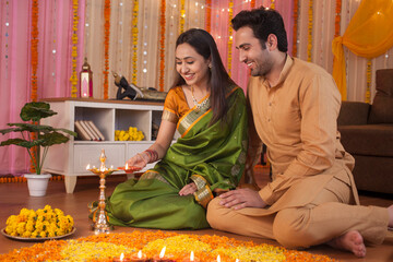 Wall Mural - Young couple decorating their house on Diwali and lighting Diyas together - Hindu festival custom. Stock image of an Indian husband-wife in traditional outfits lighting diyas and making rangoli tog...