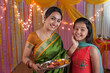 Indian mother and daughter celebrating festival and doing pooja. Mother holding a puja thali and her little daughter are standing with her. Indian attire for festival celebration