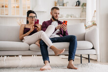 Young Caucasian Couple In Casual Sits On Couch At Home Using Phones, Searching New Apartment Via Internet. Relocation. Serious Man At Social Media. Changing Real Life For Virtual. Marriage Troubles.