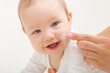 Young adult mother finger applying white moisturizing cream on baby boy cheek on light gray background. Daily care about face skin. Closeup. Front view. Happy smiling infant.