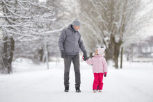Happy Smiling Little Daughter And Young Adult Father Walking On White Snow Covered Sidewalk At Park. Spending Time Together In Beautiful Cold Winter Day. Enjoying Stroll. Front View.